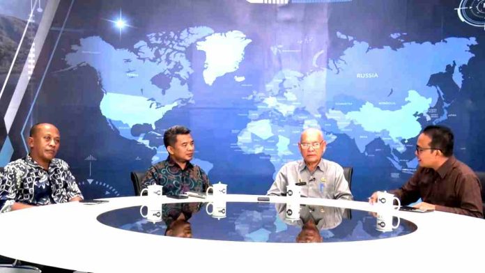 Empat orang pria duduk melingkari meja rapat putih di dalam studio TV dengan latar belakang layar besar yang menampilkan peta dunia digital berwarna biru.