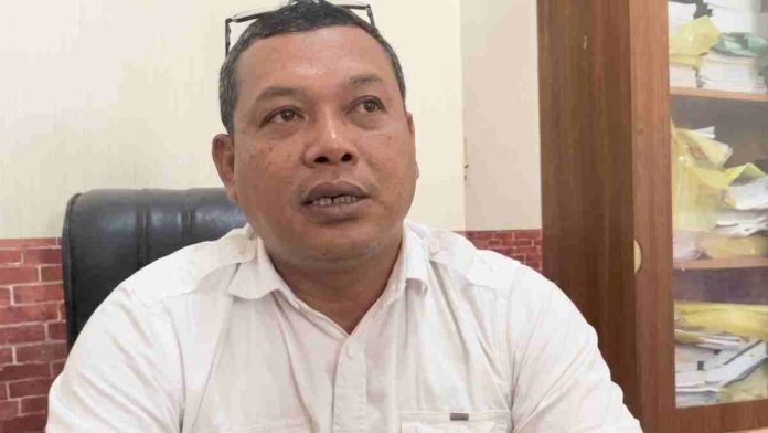 Foto close-up seorang pria paruh baya mengenakan kemeja putih dengan kacamata di atas kepala, sedang berbicara memberikan penjelasan di dalam ruang kantor.