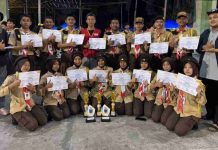 SRMA 38 Lombok Timur Borong Juara Pramuka BERAKSI VI Hamzanwadi Belasan anggota Pramuka berseragam lengkap berpose bersama memegang sertifikat dan tiga trofi juara di sebuah lapangan pada malam hari.
