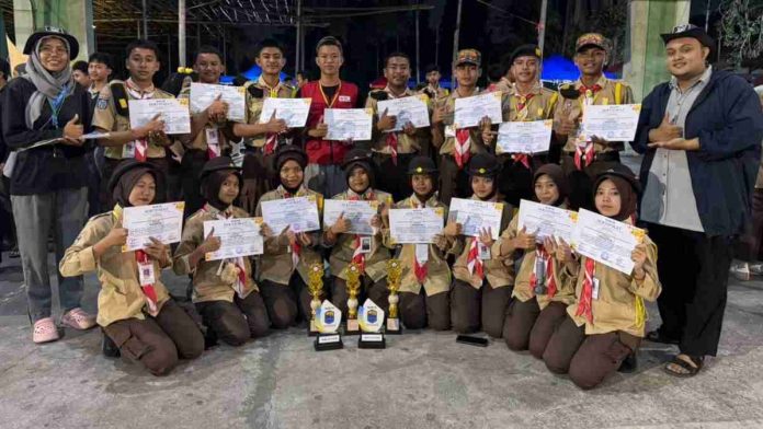 Belasan anggota Pramuka berseragam lengkap berpose bersama memegang sertifikat dan tiga trofi juara di sebuah lapangan pada malam hari.