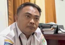 Bapenda Lombok Utara Naikkan Tarif Masuk Gili Jadi 50 Ribu Rupiah Demi PAD Foto close-up pria mengenakan seragam dinas ASN putih dengan logo Pemkab Lombok Utara di lengan, sedang memberikan keterangan resmi di depan tumpukan berkas.