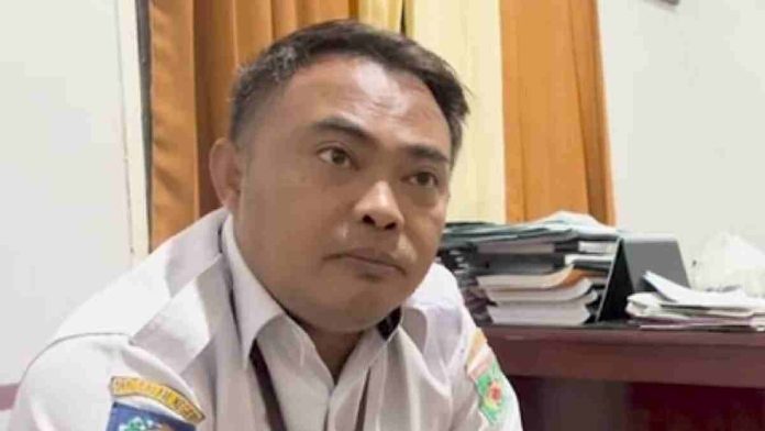 Foto close-up pria mengenakan seragam dinas ASN putih dengan logo Pemkab Lombok Utara di lengan, sedang memberikan keterangan resmi di depan tumpukan berkas.