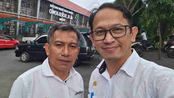 Dua pria berseragam dinas putih berdiri di area parkir dengan latar belakang gedung RSUD dr. R. Soedjono Selong yang memiliki atap merah dan fasad modern.