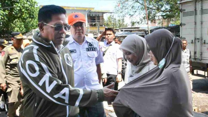 Bupati Haerul Warisin mengenakan jaket hijau gelap dan kacamata hitam sedang bersalaman dengan seorang warga di lokasi pasar, didampingi petugas BPBD berseragam putih.