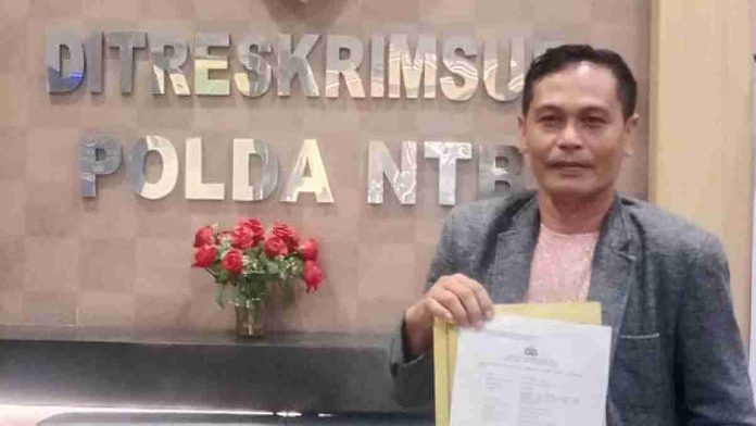 Seorang pria mengenakan jas abu-abu berdiri di depan meja resepsionis bertuliskan Ditreskrimsus Polda NTB sambil memegang berkas laporan kepolisian.
