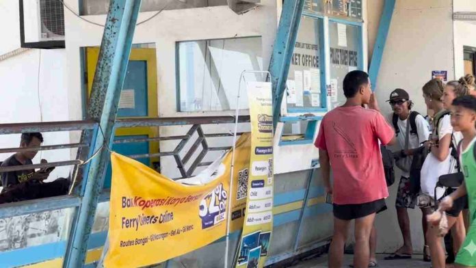 Loket pelayanan tiket pelabuhan dengan beberapa orang mengantre di depan jendela kaca, terdapat spanduk kuning informasi rute kapal di bagian depan.