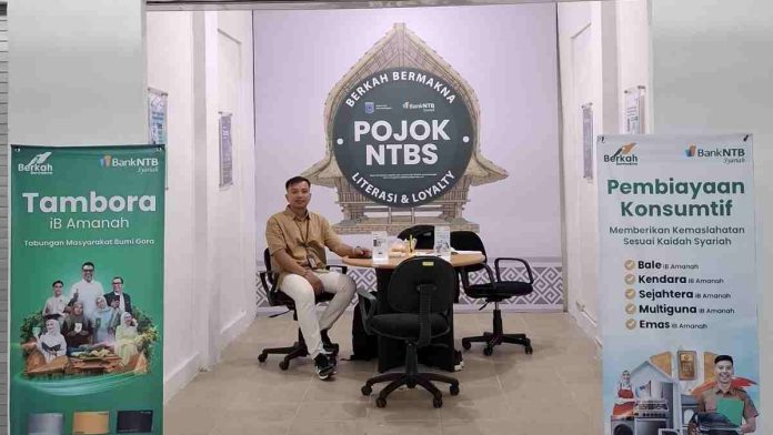 Seorang petugas pria duduk di meja layanan POJOK NTBS yang diapit oleh dua banner promosi produk tabungan dan pembiayaan syariah dalam sebuah gerai minimalis.