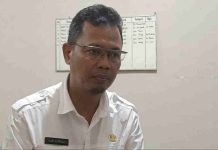 Strategi BKPSDM Lombok Timur Tambal Defisit ASN Akibat Gelombang Pensiun di Penghujung 2026 Mendatang Seorang pria berkacamata mengenakan kemeja dinas putih (pakaian ASN) dengan papan nama Hari Supriadi, tampak sedang berbicara dengan latar belakang papan jadwal kerja.