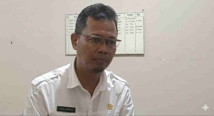 Seorang pria berkacamata mengenakan kemeja dinas putih (pakaian ASN) dengan papan nama Hari Supriadi, tampak sedang berbicara dengan latar belakang papan jadwal kerja.