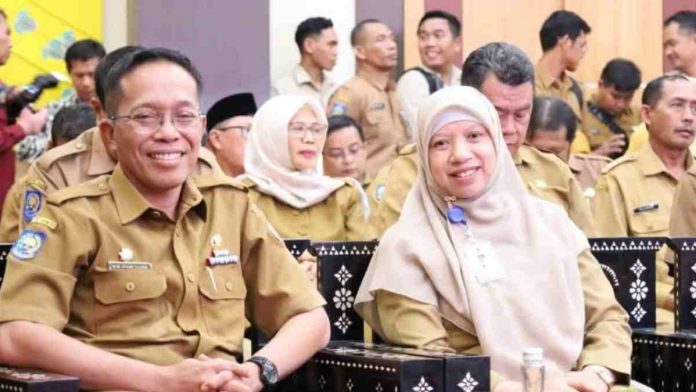 Wakil Bupati Lombok Barat Hj. Nurul Adha mengenakan jilbab merah muda dan seragam dinas cokelat, duduk berdampingan dengan pejabat pria dalam sebuah acara resmi di aula gedung.