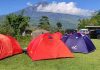 Menjaga Harmoni Rinjani melalui Tren Berkemah Keluarga di Sembalun Pemandangan area perkemahan dengan tiga tenda kubah berwarna merah dan ungu di atas padang rumput hijau, berlatar belakang Gunung Rinjani yang menjulang tinggi di bawah langit biru cerah.