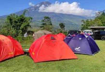 Menjaga Harmoni Rinjani melalui Tren Berkemah Keluarga di Sembalun Pemandangan area perkemahan dengan tiga tenda kubah berwarna merah dan ungu di atas padang rumput hijau, berlatar belakang Gunung Rinjani yang menjulang tinggi di bawah langit biru cerah.