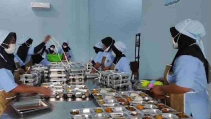 Deretan pekerja perempuan menggunakan seragam biru, apron cokelat, masker, dan penutup kepala putih sedang menyusun paket nasi kotak bergizi di atas meja stainless steel panjang.