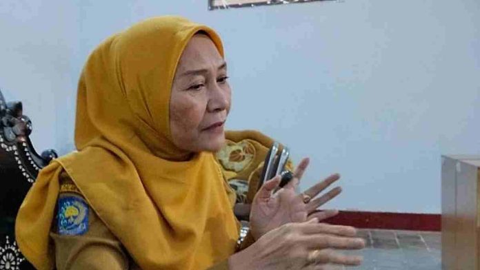 Foto close-up seorang wanita paruh baya (Kepala Dinas Sosial Siti Aminah) berhijab kuning dan seragam PNS, sedang duduk dan berbicara sambil menggerakkan kedua tangannya memberikan penjelasan.