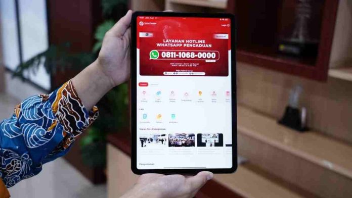 Seseorang memegang tablet yang menampilkan layar aplikasi layanan hotline WhatsApp pengaduan Kementerian ATR/BPN dengan nomor 0811-1068-0000 dan berbagai ikon menu layanan digital.