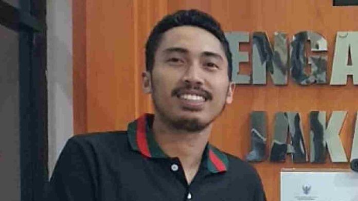 Foto close-up Firdaus Firmansyah, pria berkumis dan berjanggut tipis mengenakan kaus kerah hitam dengan garis merah-hijau, berlatar belakang dinding kayu bertuliskan 
