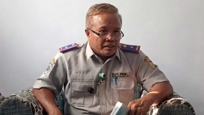 Kepala Dinas Perhubungan Safwan mengenakan seragam dinas perhubungan abu-abu dengan atribut lengkap, berkacamata, sedang memberikan keterangan di dalam ruangan.
