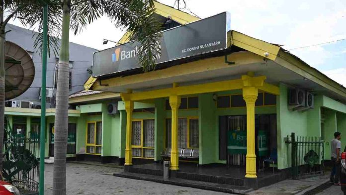 Tampak depan bangunan kantor Bank NTB Syariah KC Dompu berwarna hijau dengan aksen pilar kuning, dilengkapi papan nama besar di bagian atas serta pepohonan di halaman.