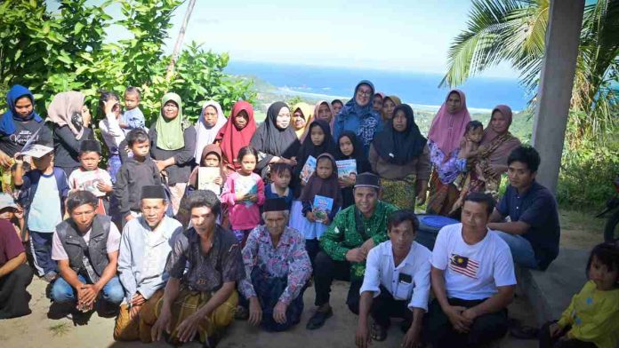 Foto bersama komunitas Tastura Mengajar dengan puluhan warga desa dan anak-anak di sebuah perbukitan dengan latar belakang pemandangan laut serta garis pantai yang biru di Lombok Tengah.