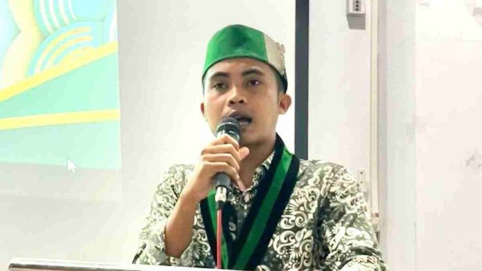 Pria berkacamata mengenakan seragam dinas perhubungan abu-abu dengan atribut lengkap di bahu, sedang duduk memberikan penjelasan dalam sebuah sesi wawancara.
