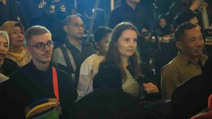 Foto jarak dekat pria berkacamata dan wanita berambut panjang sedang duduk menyimak acara peresmian di tengah kerumunan orang dalam suasana pencahayaan temaram.