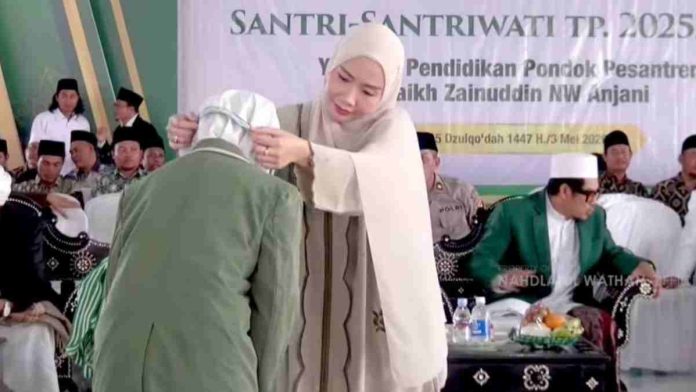 Seorang wanita berhijab krem sedang mengalungkan medali kepada seorang santri yang membungkuk, dengan latar belakang tamu undangan dan spanduk acara.