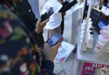 Bank NTB Syariah Sukses Terapkan QRIS Cross Border di Lima Negara Foto jarak dekat tangan seseorang memegang ponsel pintar untuk memindai kode QRIS pada sebuah cetakan kertas di depan gerai minuman UMKM.
