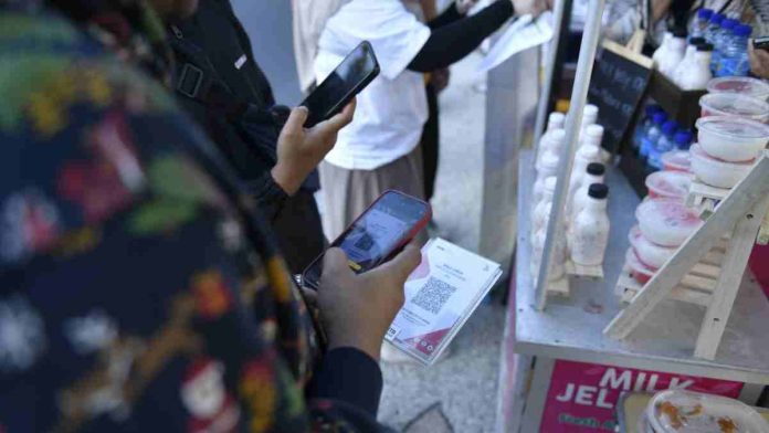 Foto jarak dekat tangan seseorang memegang ponsel pintar untuk memindai kode QRIS pada sebuah cetakan kertas di depan gerai minuman UMKM.