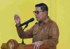 Pasar Pringgabaya Bakal Dikembalikan Seperti Semula, Bupati Lotim: Plungguh Sabar Bupati Lombok Timur H. Haerul Warisin mengenakan seragam cokelat dan kacamata hitam sedang berpidato di balik podium dengan latar belakang dinding kuning.