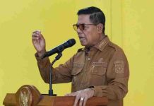 Pasar Pringgabaya Bakal Dikembalikan Seperti Semula, Bupati Lotim: Plungguh Sabar Bupati Lombok Timur H. Haerul Warisin mengenakan seragam cokelat dan kacamata hitam sedang berpidato di balik podium dengan latar belakang dinding kuning.