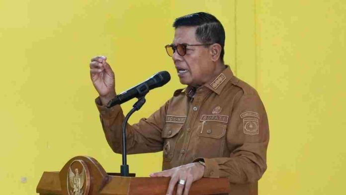 Bupati Lombok Timur H. Haerul Warisin mengenakan seragam cokelat dan kacamata hitam sedang berpidato di balik podium dengan latar belakang dinding kuning.