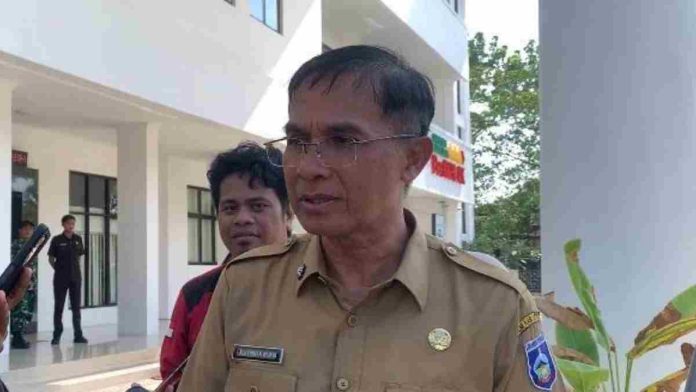 Foto Sekretaris Daerah Lombok Tengah mengenakan seragam dinas cokelat dan kacamata, berdiri memberikan penjelasan kepada awak media di depan gedung.
