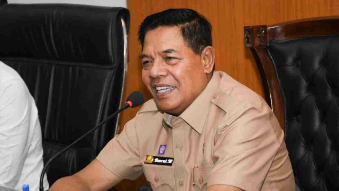 Bupati Lombok Timur H. Haerul Warisin mengenakan seragam dinas cokelat, sedang berbicara dengan ekspresi serius di depan mikrofon dalam sebuah pertemuan resmi.
