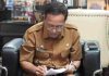 Bidik Hibah Bank Dunia, Pemkab Lombok Timur Siapkan Lahan TPST Terpadu Sekda Lombok Timur H. Muhammad Juaini Taofik mengenakan seragam dinas cokelat dan kacamata, tampak fokus menulis atau menandatangani dokumen di atas pangkuannya.