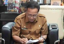 Bidik Hibah Bank Dunia, Pemkab Lombok Timur Siapkan Lahan TPST Terpadu Sekda Lombok Timur H. Muhammad Juaini Taofik mengenakan seragam dinas cokelat dan kacamata, tampak fokus menulis atau menandatangani dokumen di atas pangkuannya.