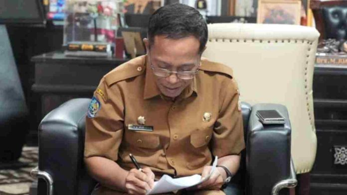 Sekda Lombok Timur H. Muhammad Juaini Taofik mengenakan seragam dinas cokelat dan kacamata, tampak fokus menulis atau menandatangani dokumen di atas pangkuannya.