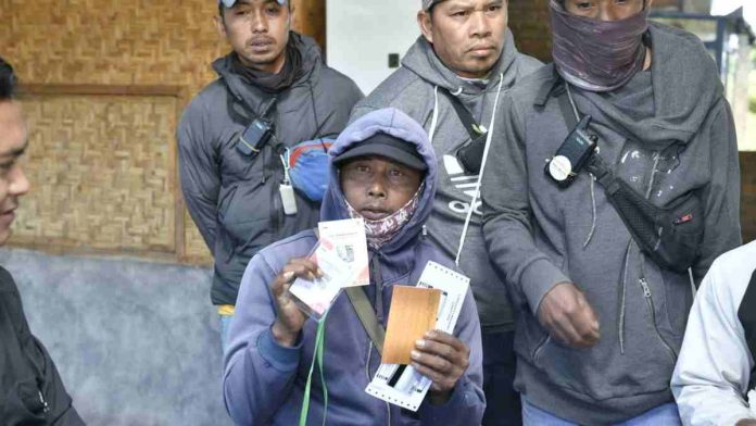 Seorang pria mengenakan jaket hoodie ungu menunjukkan kartu QRIS dan dokumen aktivasi merchant di depan rekan-rekannya sesama penyedia jasa ojek wisata.