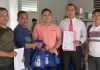 Disdag Lotim Tegaskan HPS Sembako Sesuai Survei Pasar dan Kawalan APH Seorang pria berbaju putih berbicara memegang mikrofon dengan latar belakang backdrop biru besar yang menampilkan foto tokoh nasional.