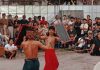 Lestarikan Budaya, Bajang Loyok Metangi Gelar Peresean Skala Lombok Dua orang pria (pepadu) bertelanjang dada mengenakan kain tradisional sedang bertarung Peresean menggunakan rotan dan perisai di tengah lapangan tanah. Di latar belakang, tampak kerumunan warga lokal dan turis asing menyaksikan pertandingan dengan antusias.
