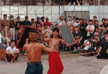 Lestarikan Budaya, Bajang Loyok Metangi Gelar Peresean Skala Lombok Dua orang pria (pepadu) bertelanjang dada mengenakan kain tradisional sedang bertarung Peresean menggunakan rotan dan perisai di tengah lapangan tanah. Di latar belakang, tampak kerumunan warga lokal dan turis asing menyaksikan pertandingan dengan antusias.