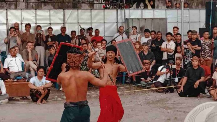 Dua orang pria (pepadu) bertelanjang dada mengenakan kain tradisional sedang bertarung Peresean menggunakan rotan dan perisai di tengah lapangan tanah. Di latar belakang, tampak kerumunan warga lokal dan turis asing menyaksikan pertandingan dengan antusias.